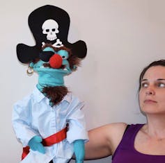 A pirate puppet who looks crochety is seen on the hand of a pupetteer.