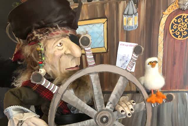 A pirate puppet holds a ship's steering wheel and a seagull puppet is perched on it. 