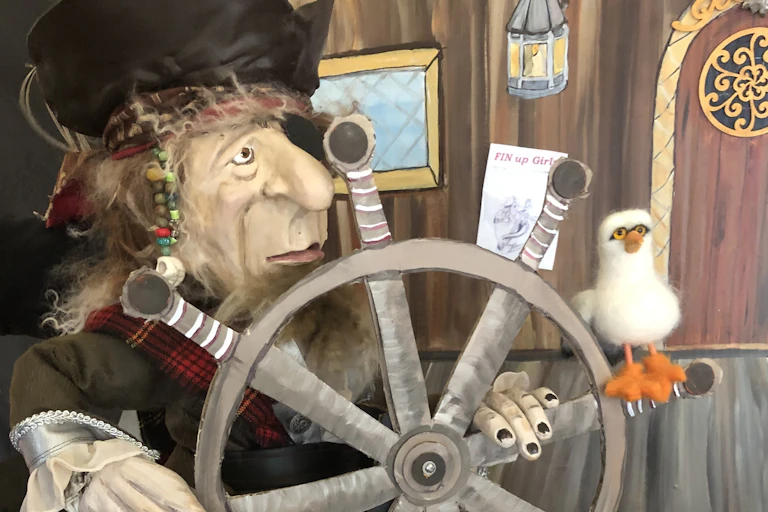 A pirate puppet holds a ship's steering wheel and a seagull puppet is perched on it.