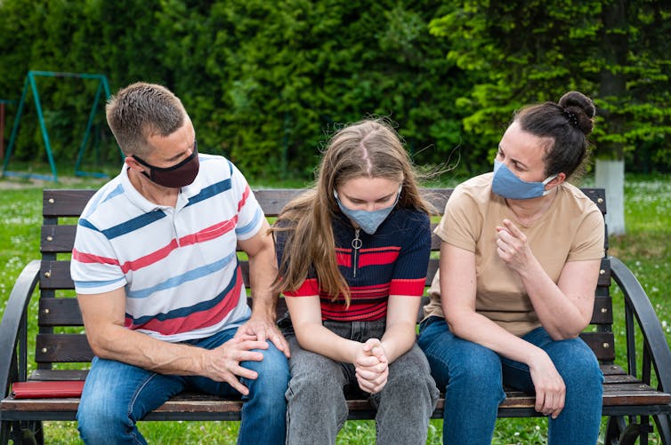 Parents and their child sitting on a park bench, wearing masks.