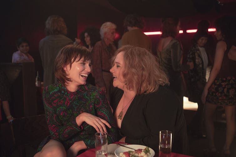 Movie still, Helen sits in Lillian's lap at a bar as they laugh.