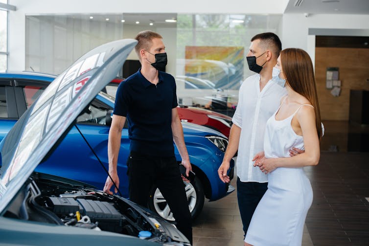 Young couple in masks talk to a salesman in front of a car.