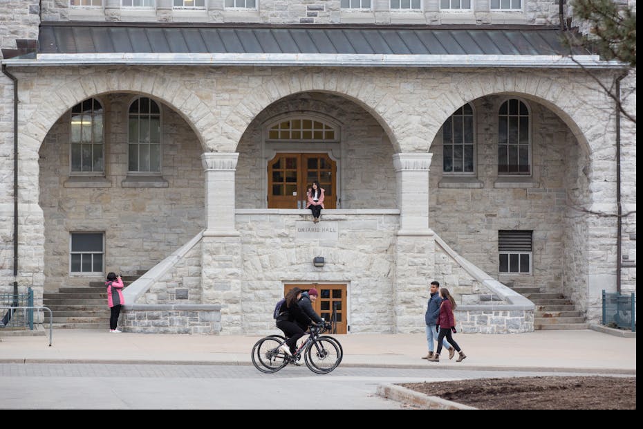 A student is seen riding a bicycle in front of a university building while while others stroll past.