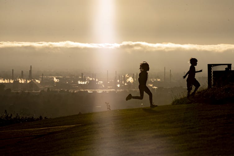 Two children running on a hill, oil refinery in background.
