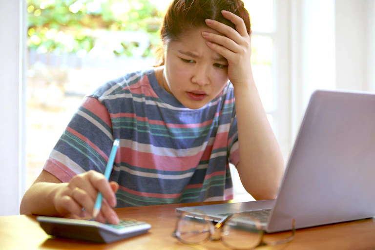 A young woman sits on a desk with her laptop open while typing on a calculator.