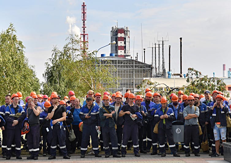 Belarusian workers in orange hardhats stand with arms crossed in front of a factory