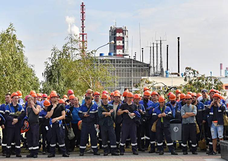 Belarusian workers in orange hardhats stand with arms crossed in front of a factory