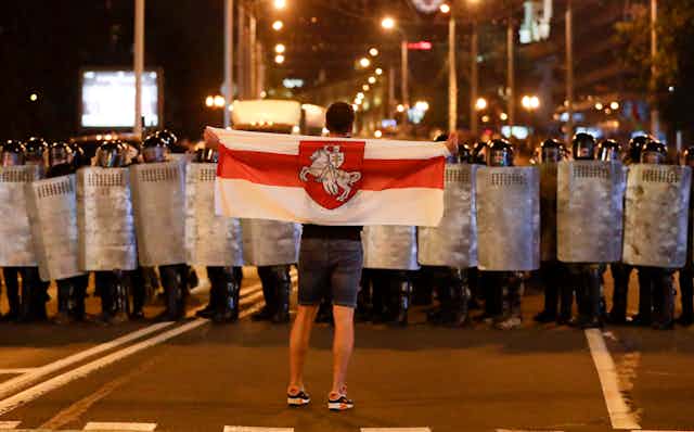 A sole man holding the Belarussian flag faces a line of police in riot gear