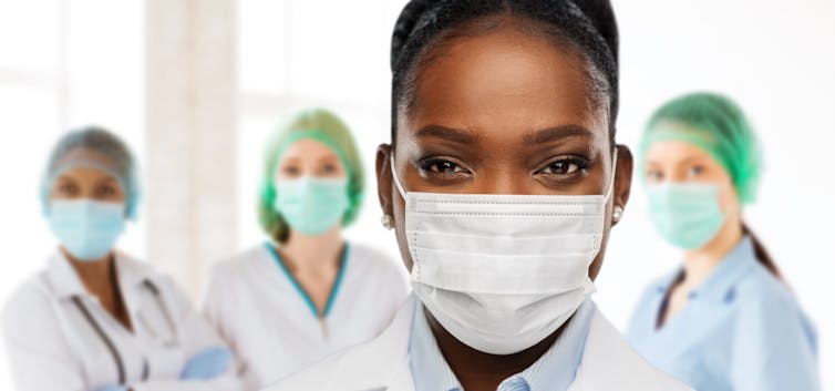 Close-up of a woman's face in a mask in the foreground, with three nurses in masks and PPE in the background.