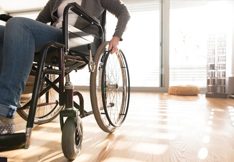 Woman sits in a wheelchair indoors. Her head is cut off.