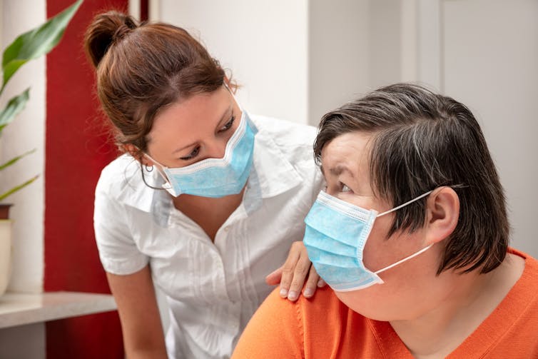 A carer has her hand on the shoulder of a woman with disability. Both are wearing masks.