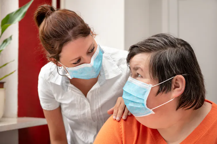 A carer has her hand on the shoulder of a woman with disability. Both are wearing masks.
