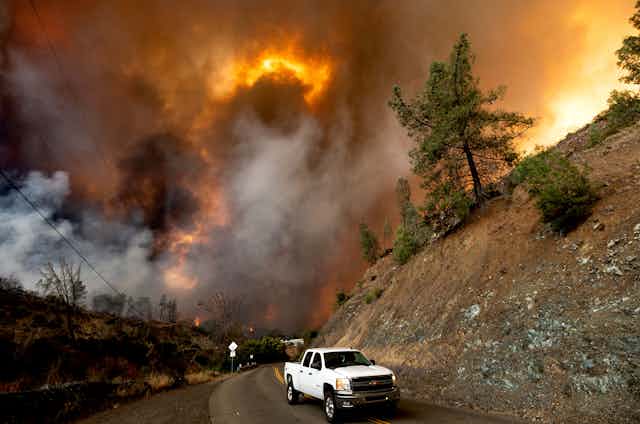 A truck drives out a valley where a wildfire burns.
