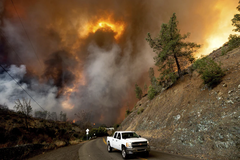 A truck drives out a valley where a wildfire burns.