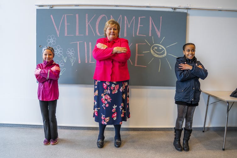 The Norwegian prime minister stands in front of a blackboard with two children, all with arms folded.