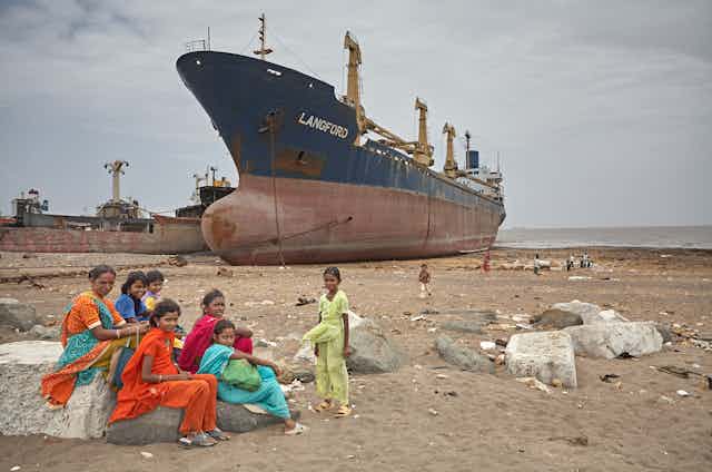 A group of women sit on a beach in front of a cargo ship.