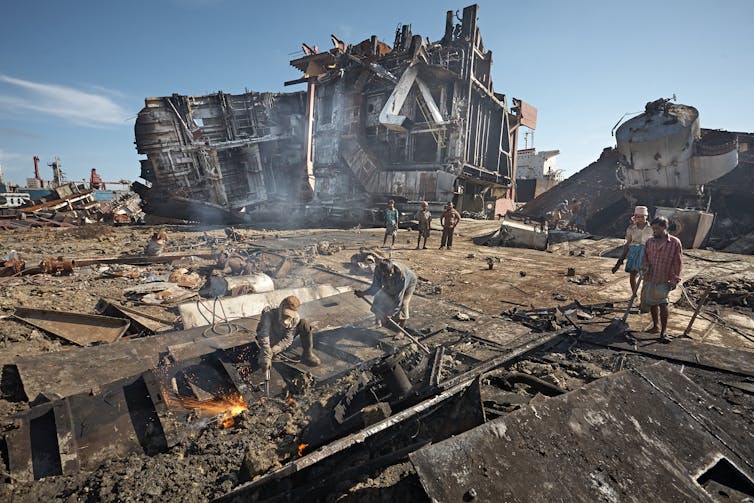 People dismantling parts of a ship, large ship on its side in the background.