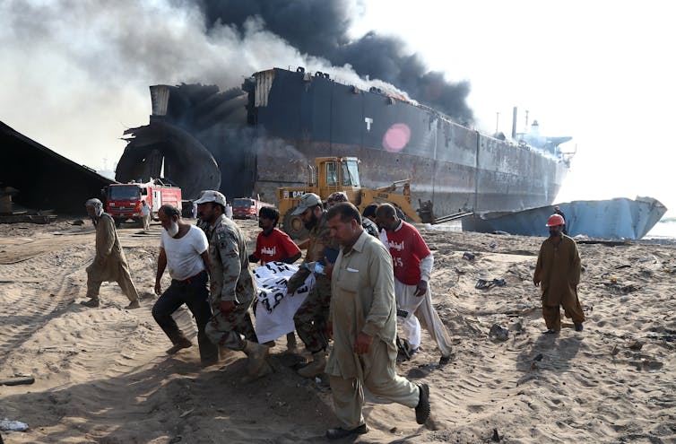 Victim carried across beach on stretcher, large ship in background with smoke.