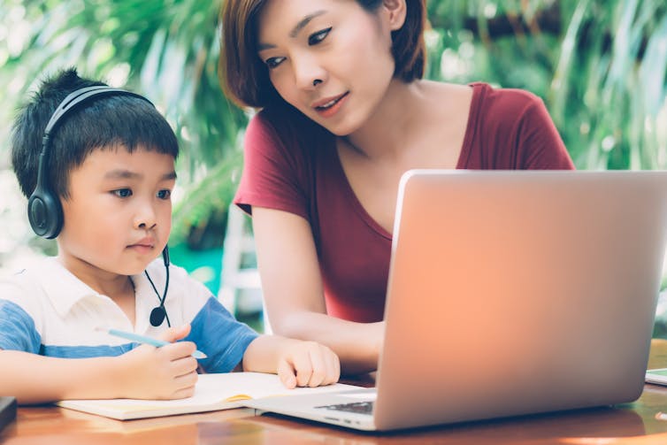 A young child sits outdoors in front of a laptop wearing headphones, while his mother, sitting next to him, leans towards him.