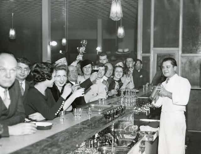 Fotografía en blanco y negro de hombres y mujeres bebiendo y brindado en una barra de bar, con el barman haciendo un cóctel al otro lado de la barra.