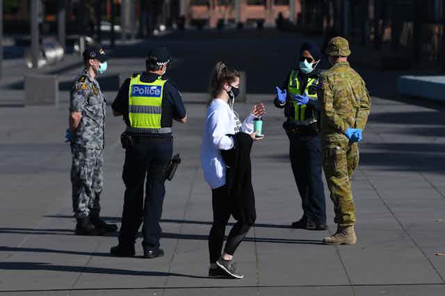 A lady wearing a mask, walks passed military and police officers, in metropolitan Melbourne