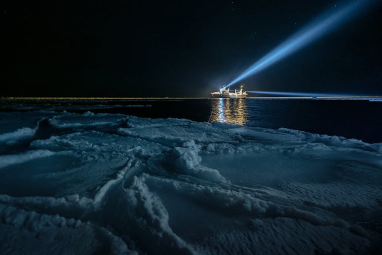 Seen from a sea ice floe, a large ship on the horizon beams white light into the sky.