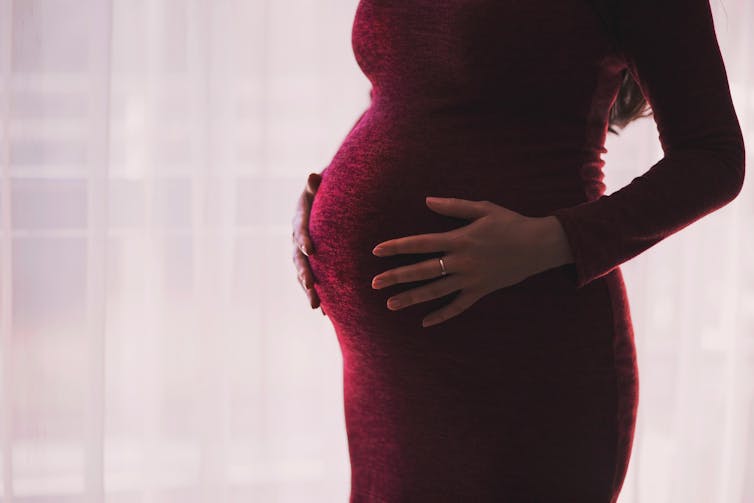 A pregnant woman from the shoulders down, wearing a red dress, with her hands on her belly.