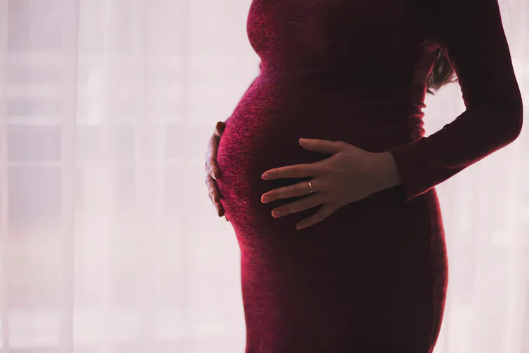 A pregnant woman from the shoulders down, wearing a red dress, with her hands on her belly.