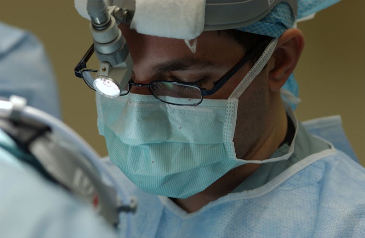 Close-up of a male surgeon's face looking down, wearing a surgical mask and cap, glasses and a forehead-mounted light.