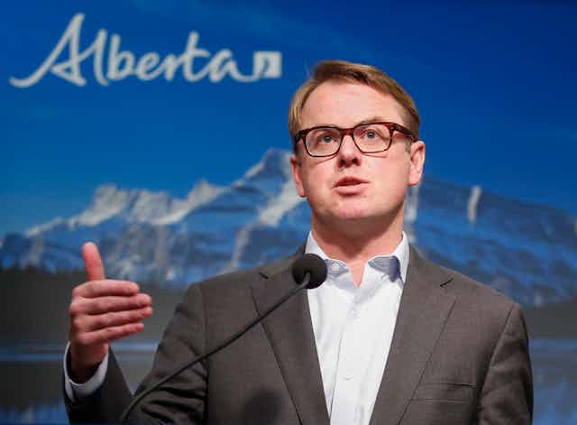 Alberta Minister of Health Tyler Shandro standing at a podium in front of a backdrop of mountains and the word 'Alberta'