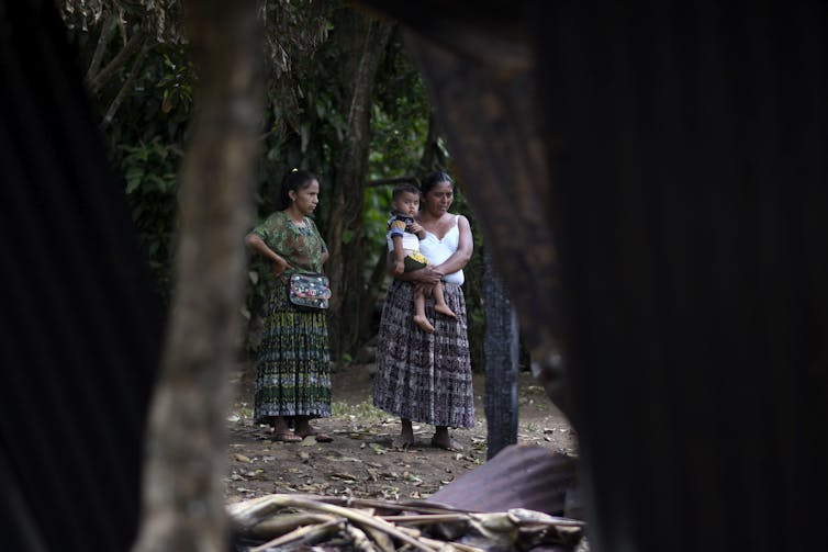 Two women and a child in traditional Indigenous clothing look at a crime scene where a home was burned