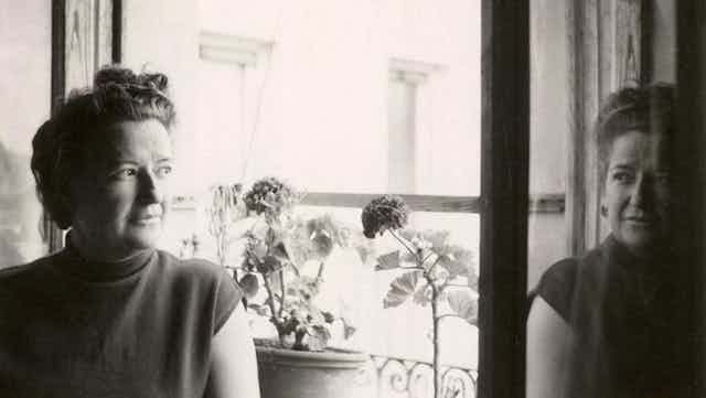 Fotografía en blanco y negro de Carmen Conde junto a una ventana con flores y el reflejo de su rostro en el cristal.