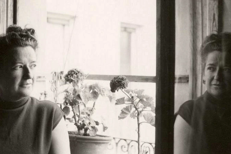 Fotografía en blanco y negro de Carmen Conde junto a una ventana con flores y el reflejo de su rostro en el cristal.
