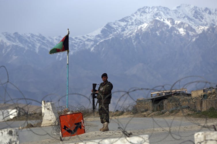 Afghan soldier in fatigues guards a checkpoint, with snow-capped mountains and an Afghan flag in the background