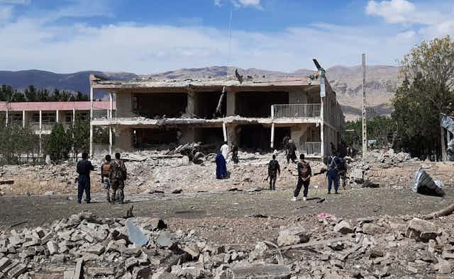 Soldiers and civilians stand in the rubble of a collapsed building with mountains in the background