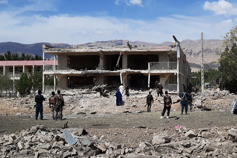 Soldiers and civilians stand in the rubble of a collapsed building with mountains in the background