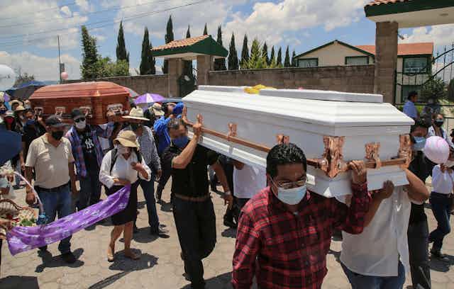Men in face masks carry two coffins, one white and one mahogany