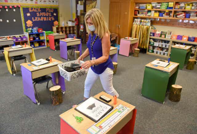 An elementary school teacher prepares her classroom for students, with desks spaced several feet apart.
