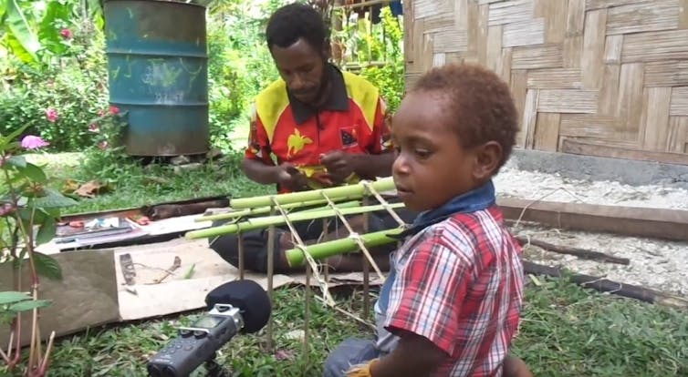 A young boy looks at a voice recorder while his father works in the background.