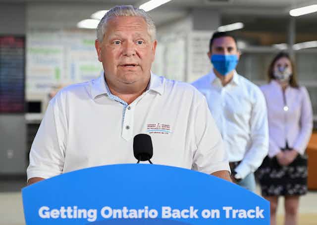 Doug Ford speaks into a microphone in front of a podium that reads Getting Ontario Back on Track, with two masked officials standing behind him.