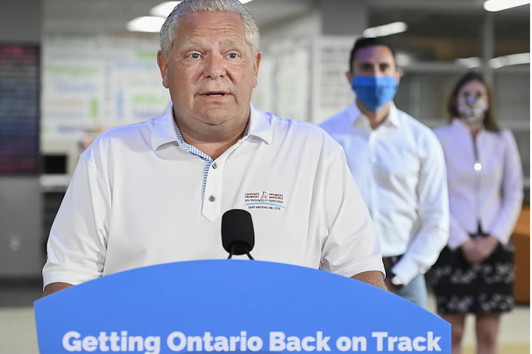Doug Ford speaks into a microphone in front of a podium that reads Getting Ontario Back on Track, with two masked officials standing behind him.