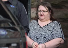 A woman in glasses and handcuffs is led to a police cruiser.