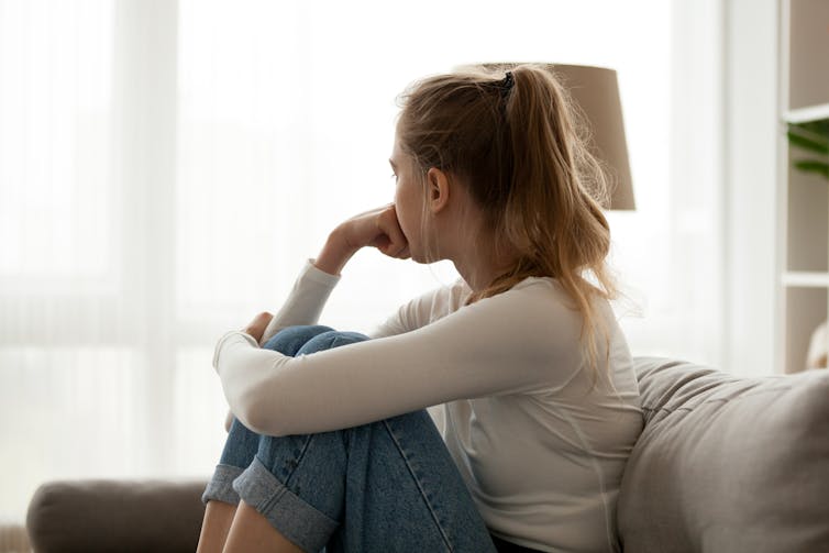 A side view of a girl on the couch, with knees to her chest and chin on hand looking away from camera.