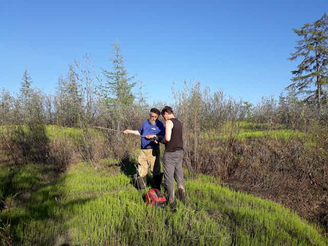 Two men stand on a grassy bank with some technical equipment.