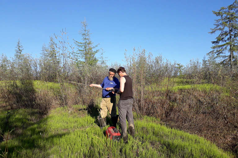 Two men stand on a grassy bank with some technical equipment.