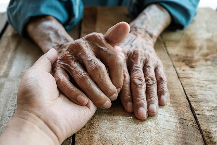 One hand reaches out to hold a pair of wrinkled hands on a wooden table.