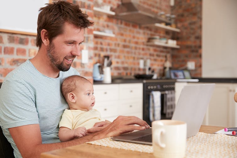 A man typing on a laptop with his baby sitting on his knee.