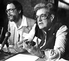 A woman wearing glasses speaks into a microphone in a black-and-white photo.