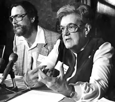 A woman wearing glasses speaks into a microphone in a black-and-white photo.