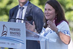 A woman with dark shoulder-length hair speaks into a microphone behind a podium that reads Premiers Ministres.